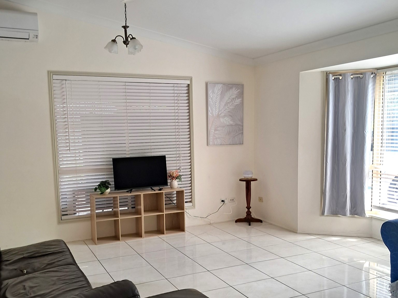 Furnished living room in the Kanda Frenchville SIL Home featuring white tiled floor, air conditioning, white Venetian blinds, and a TV on a wooden stand.
