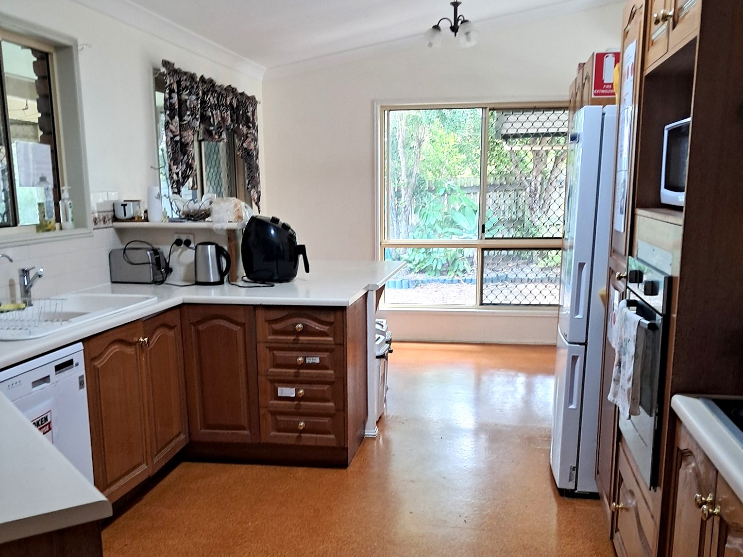 Traditional kitchen in the Kanda Frenchville SIL Home with wooden cabinetry, white benchtops, orange/brown floor, and a view of the screened outdoor area.