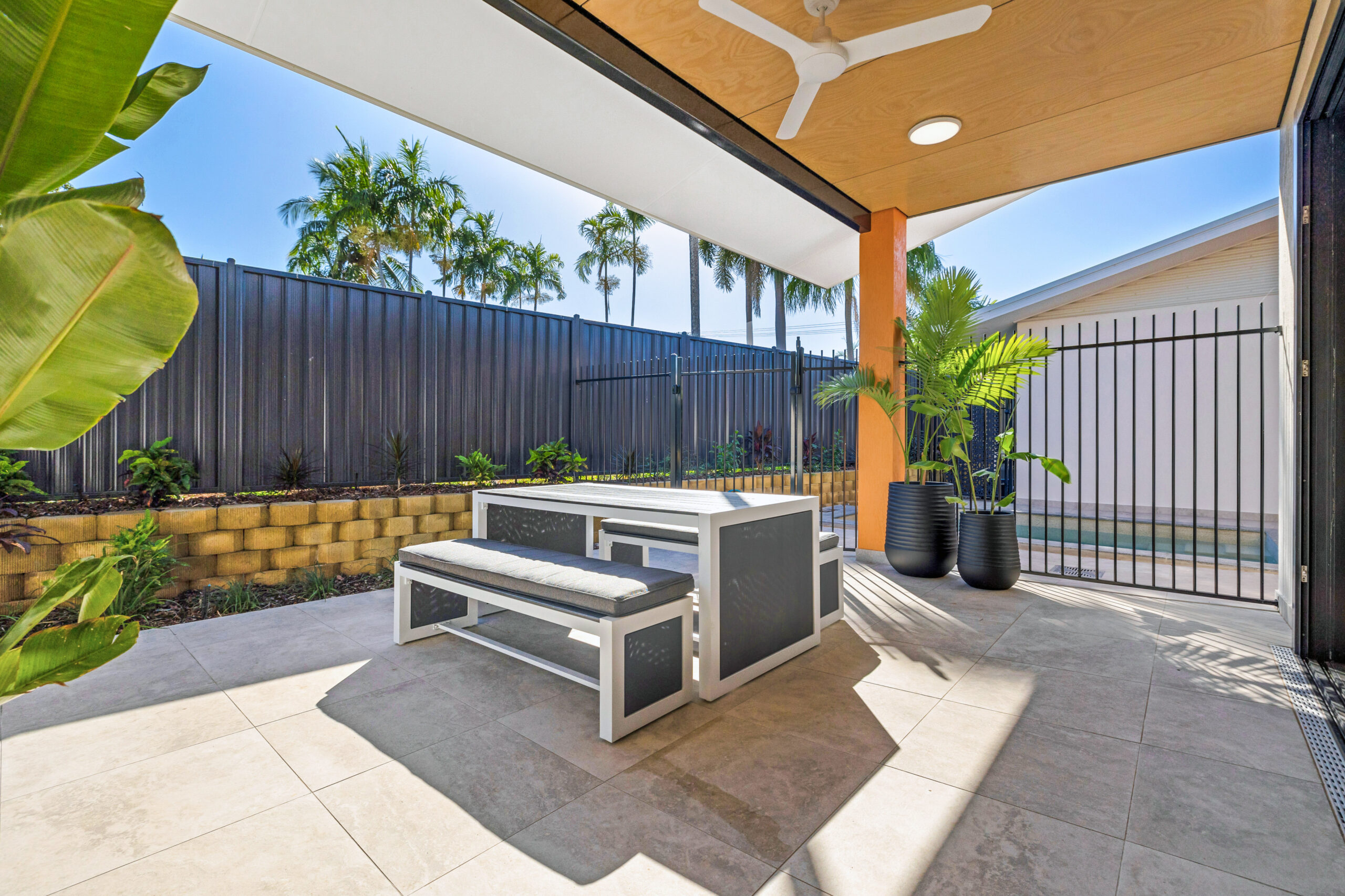 SDA home exterior patio, featuring outdoor bench seats and table. Two plants in black pots sit in front of the pool fence.