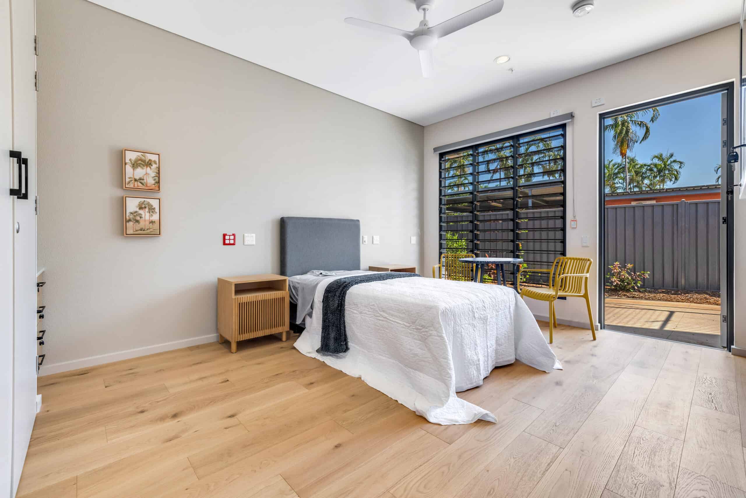 SDA home, bedroom, featuring a single bed, light wooden bedside table and a yellow chair. The bedroom door opens to a small garden space.